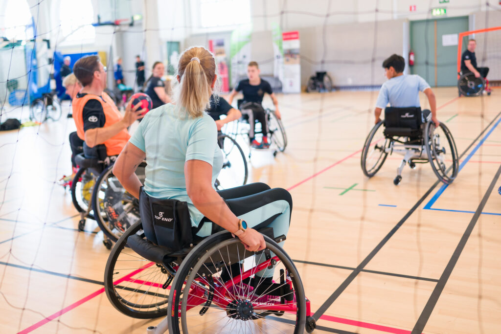 I förgrunden en kvinna i rullstol vänd bort mot ett antal personer i rullstolar som spelar handboll i en idrottshall.
